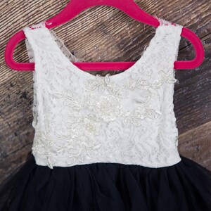 Puede incluir: Un vestido blanco sin mangas de encaje con una falda de tul negro. El vestido est&aacute; en una percha rosa.