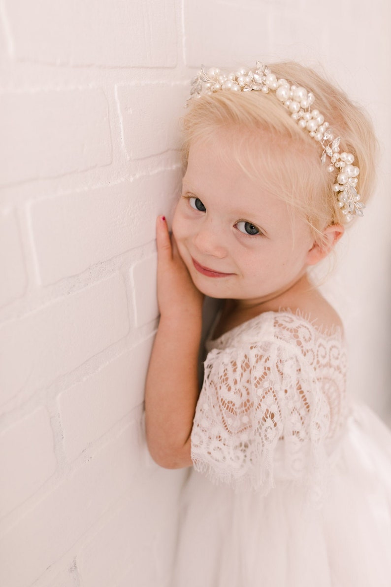 May include: A young girl with blonde hair wearing a white lace dress and a pearl and rhinestone headband. She is smiling and looking at the camera.