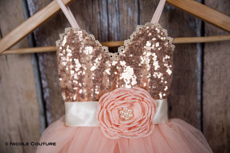 May include: A blush pink tulle dress with a rose gold sequin bodice and a white satin sash. The dress has a large blush pink flower with pearl accents on the front.