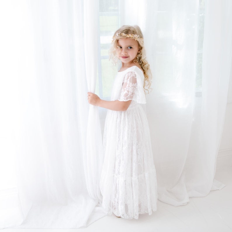 May include: A young girl wearing a white lace dress with a gold crown, standing by a white curtain and looking at the camera.