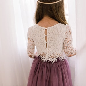 May include: A white lace top with a button-up back and 3/4 sleeves, paired with a dusty purple tulle skirt.