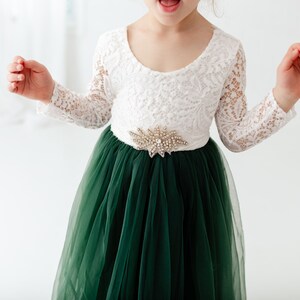 K&ouml;nnte beinhalten: Ein wei&szlig;es Spitzenkleid mit langen &Auml;rmeln, einem gr&uuml;nen T&uuml;llrock und einem G&uuml;rtel mit Schmucksteinen.