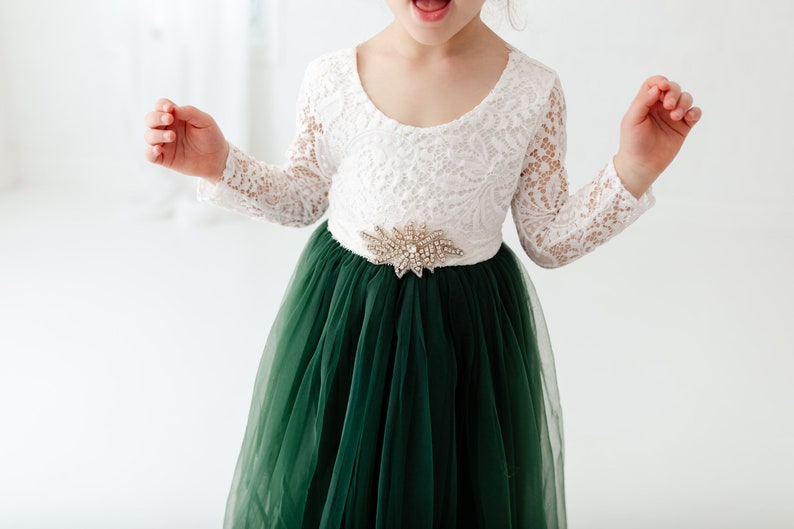 K&ouml;nnte beinhalten: Ein wei&szlig;es Spitzenkleid mit langen &Auml;rmeln, einem gr&uuml;nen T&uuml;llrock und einem G&uuml;rtel mit Schmucksteinen.