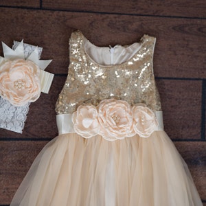 Op de afbeelding: Een gouden paillettenjurk met een tule rok en een satijnen band met perzikkleurige bloemen van stof. Een bijpassend hoofdband met een perzikkleurige bloem van stof en kant.
