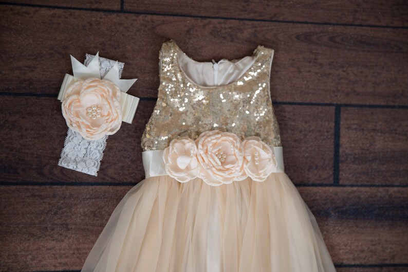 Op de afbeelding: Een gouden paillettenjurk met een tule rok en een satijnen band met perzikkleurige bloemen van stof. Een bijpassend hoofdband met een perzikkleurige bloem van stof en kant.