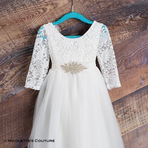 Puede incluir: Vestido blanco de encaje y tul con mangas largas y un cintur&oacute;n brillante. El vestido tiene un escote redondo y una falda completa.