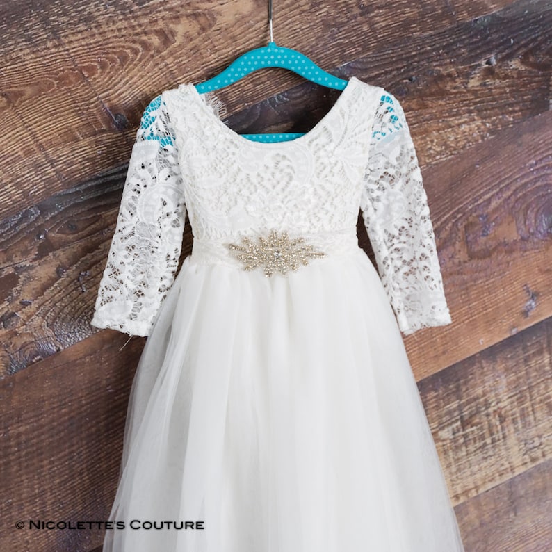 Puede incluir: Vestido blanco de encaje y tul con mangas largas y un cintur&oacute;n brillante. El vestido tiene un escote redondo y una falda completa.