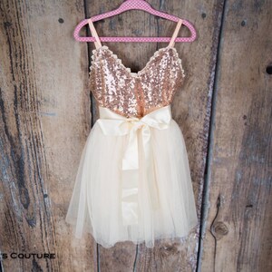 May include: A gold and ivory tulle dress with a sequined bodice and a satin bow. The dress is hanging on a pink hanger against a wooden background.