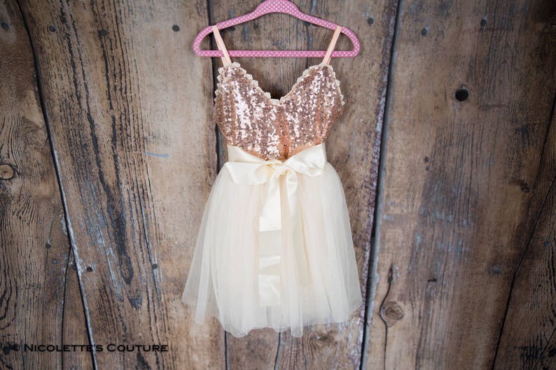 May include: A gold and ivory tulle dress with a sequined bodice and a satin bow. The dress is hanging on a pink hanger against a wooden background.