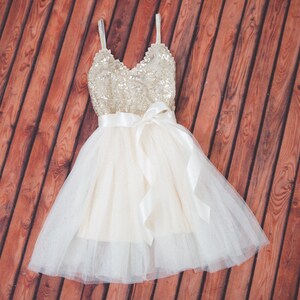 May include: A sleeveless dress featuring a gold sequined bodice with a V-neckline and thin straps. The waist is accented with a wide white satin ribbon tied in a bow. The skirt consists of multiple layers of white tulle over a solid base fabric.
