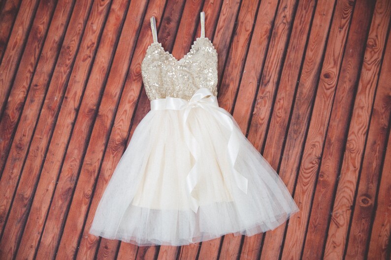 May include: A sleeveless dress featuring a gold sequined bodice with a V-neckline and thin straps. The waist is accented with a wide white satin ribbon tied in a bow. The skirt consists of multiple layers of white tulle over a solid base fabric.