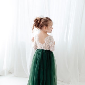 Pu&ograve; includere: Un abito lungo fino ai piedi con corpetto in pizzo bianco a maniche lunghe e scollo profondo sulla schiena, abbinato a un'ampia gonna in tulle verde scuro. Il capo presenta una parte superiore aderente e una gonna voluminosa a pi&ugrave; strati.