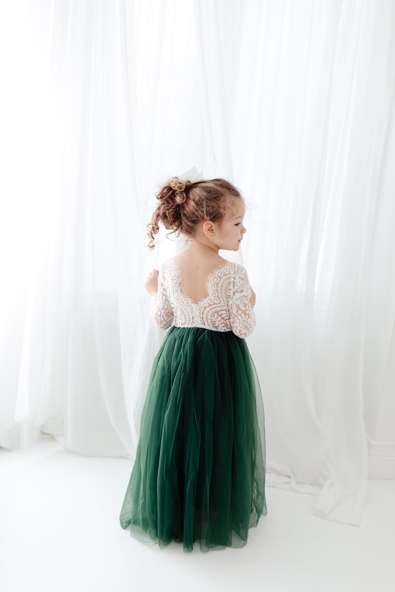 Pu&ograve; includere: Un abito lungo fino ai piedi con corpetto in pizzo bianco a maniche lunghe e scollo profondo sulla schiena, abbinato a un'ampia gonna in tulle verde scuro. Il capo presenta una parte superiore aderente e una gonna voluminosa a pi&ugrave; strati.