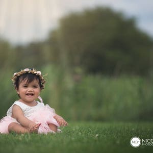 May include: A young girl wearing a pink dress and a flower crown sits on a grassy field. She is smiling and looking at the camera.