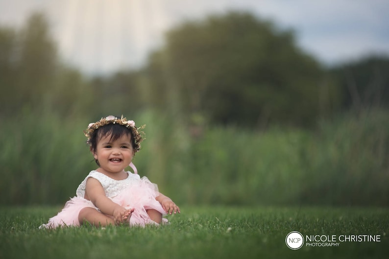 May include: A young girl wearing a pink dress and a flower crown sits on a grassy field. She is smiling and looking at the camera.