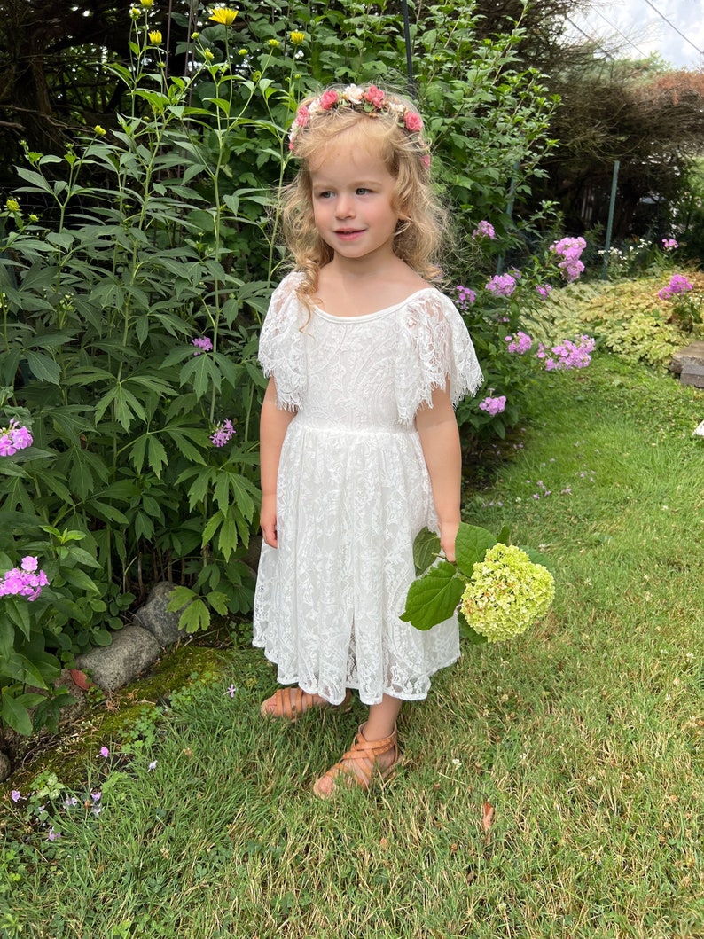 May include: A young girl wearing a white lace dress with a floral crown and holding a bouquet of white flowers. She is standing in a grassy area with green foliage and flowers.