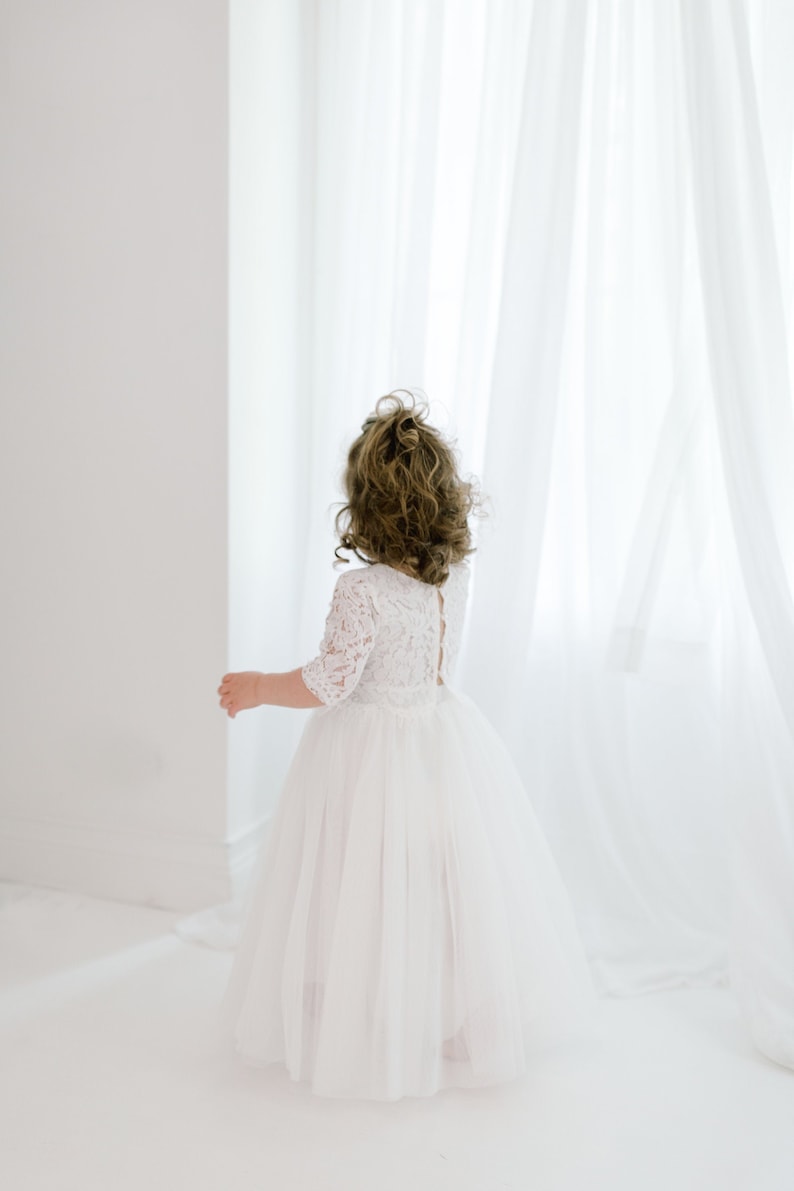 May include: A young girl wearing a white lace dress with a tulle skirt. She is standing in front of a white curtain.