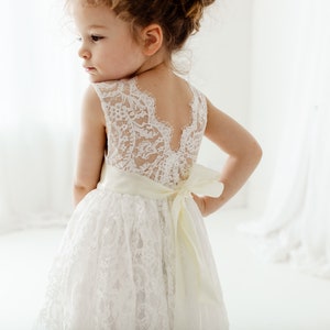 May include: A white sleeveless dress featuring an intricate floral lace overlay on the bodice and skirt. The back of the dress has a deep V-shaped cutout and a wide, smooth cream-colored satin ribbon tied into a bow at the waistline.
