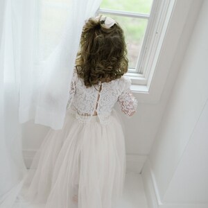 May include: A young girl wearing a white lace top and tulle skirt stands by a window, looking outside. The outfit is a light cream color, and the girl's hair is curly. A white curtain is to the left of the girl.