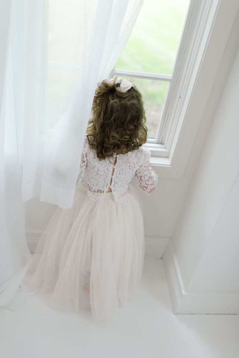 May include: A young girl wearing a white lace top and tulle skirt stands by a window, looking outside. The outfit is a light cream color, and the girl's hair is curly. A white curtain is to the left of the girl.