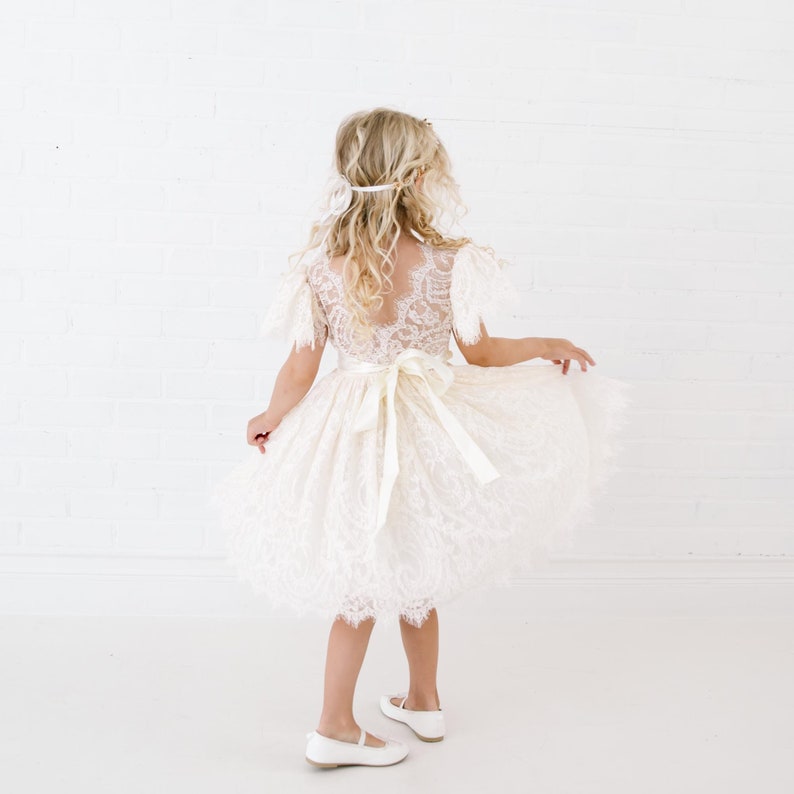 May include: A cream-colored lace dress featuring short flutter sleeves, a deep V-shaped back, and a wide satin ribbon tied into a bow at the waist. The skirt is full and layered with intricate floral lace patterns, paired with simple white flat shoes.