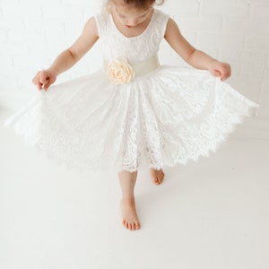 Pode incluir: Uma menina pequena vestindo um vestido branco de renda com uma faixa de cetim e um detalhe floral. O vestido tem uma saia cheia e &eacute; perfeito para uma ocasi&atilde;o especial.