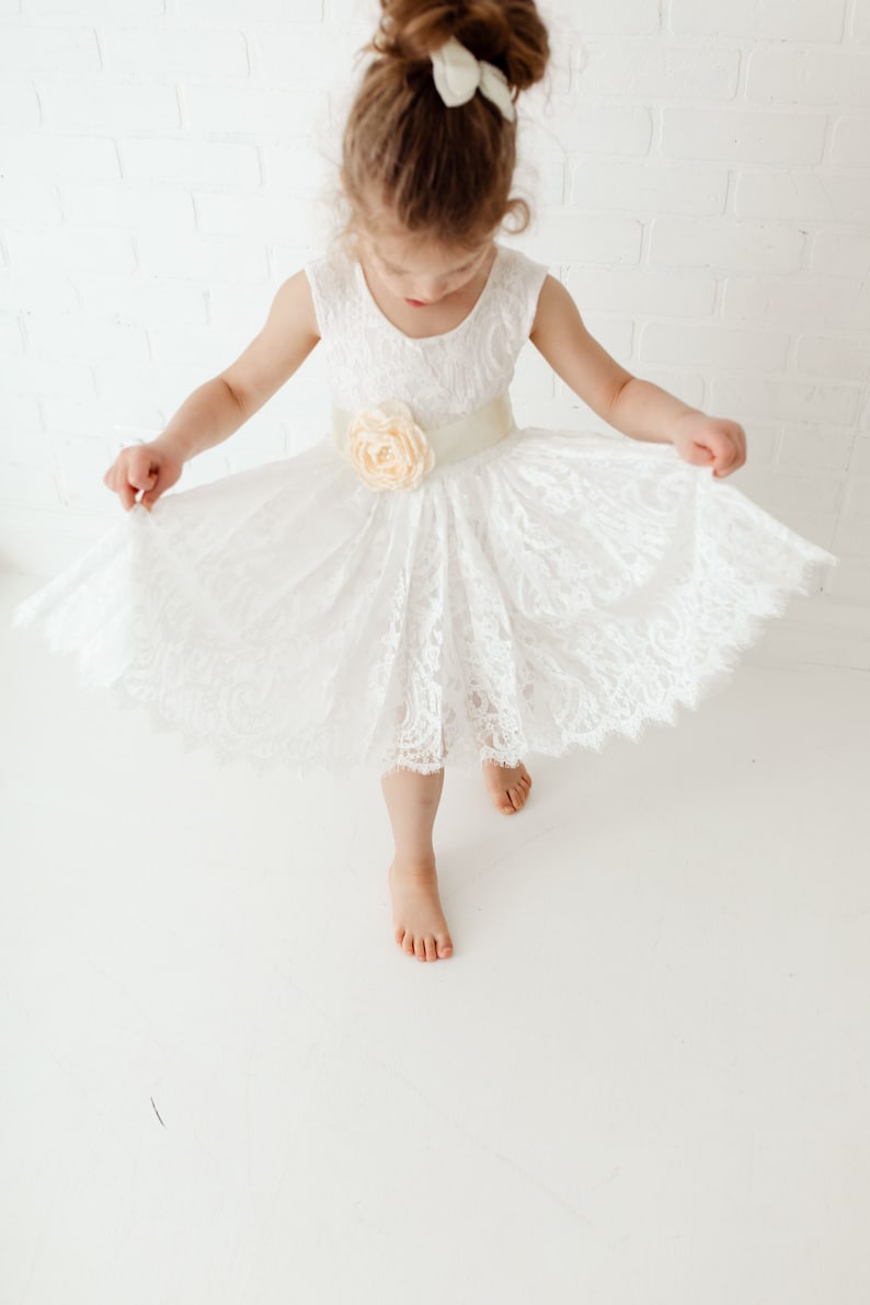 Pode incluir: Uma menina pequena vestindo um vestido branco de renda com uma faixa de cetim e um detalhe floral. O vestido tem uma saia cheia e &eacute; perfeito para uma ocasi&atilde;o especial.
