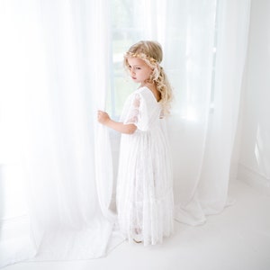 May include: A young girl wearing a white lace dress with a gold floral crown stands by a window, looking out. The dress has a ruffled skirt and short sleeves.