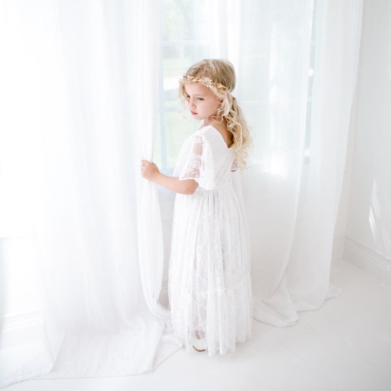 May include: A young girl wearing a white lace dress with a gold floral crown stands by a window, looking out. The dress has a ruffled skirt and short sleeves.