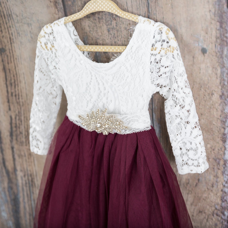 May include: A long-sleeved dress featuring a white lace bodice with a scoop neckline and a deep burgundy tulle skirt. A decorative gold-toned rhinestone embellishment is centered at the waistline where the lace bodice meets the tulle skirt.