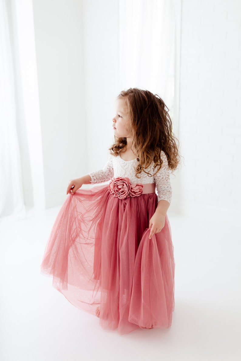 May include: A young girl wearing a white lace top with a pink tulle skirt and a pink flower sash. The skirt is flowing and the girl is looking to the side.