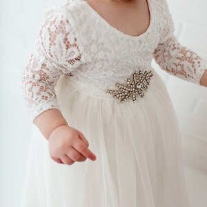 Pu&ograve; includere: Un abito di tulle bianco con maniche di pizzo e un ornamento di gioielli in vita.