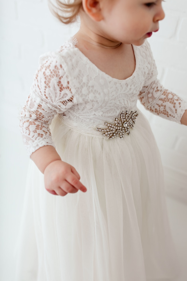 Pu&ograve; includere: Un abito di tulle bianco con maniche di pizzo e un ornamento di gioielli in vita.