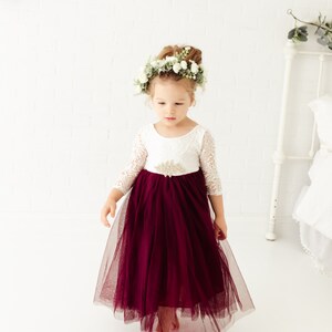 May include: A young girl wearing a white lace dress with a burgundy tulle skirt and a flower crown. She is standing on a white background.