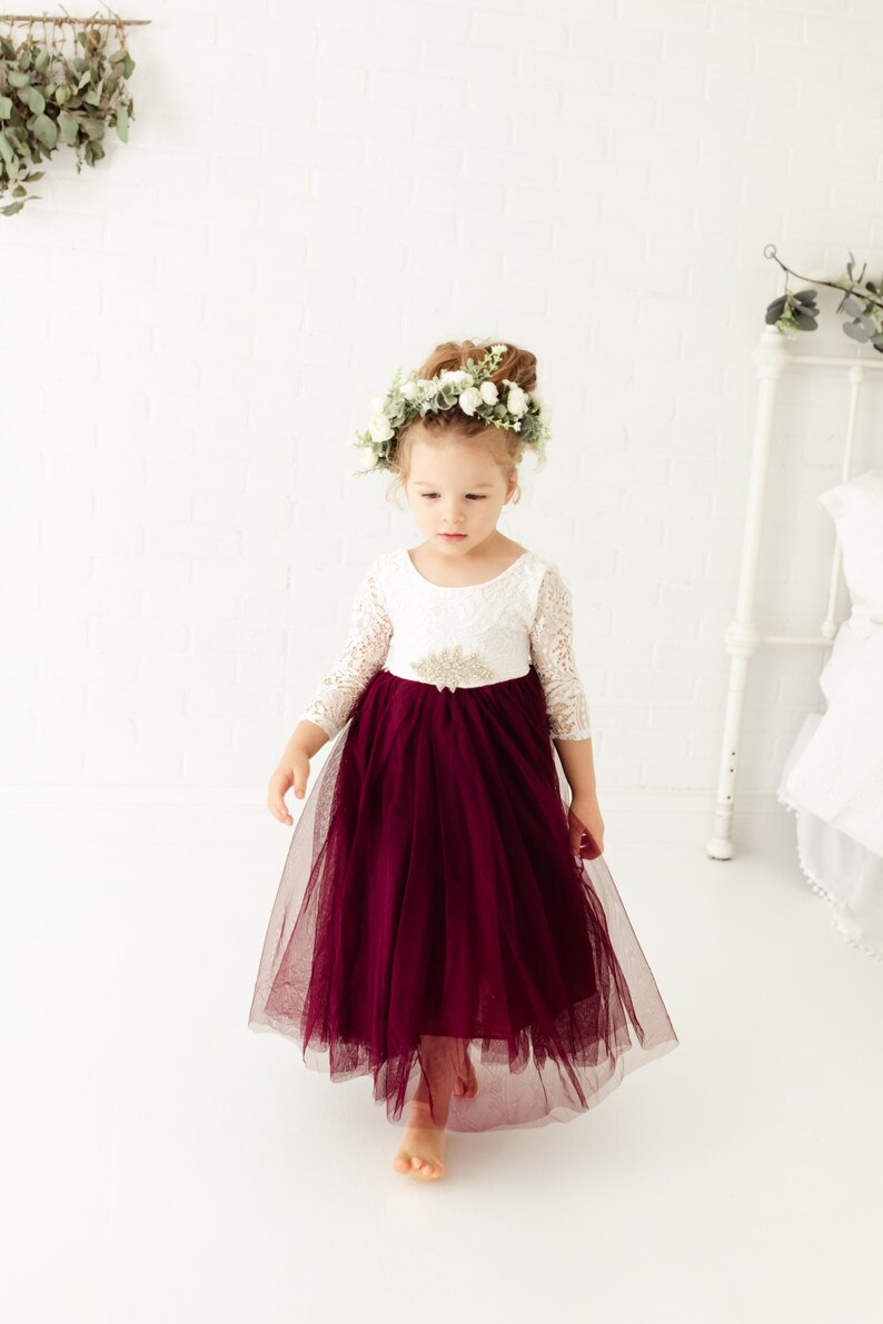 May include: A young girl wearing a white lace dress with a burgundy tulle skirt and a flower crown. She is standing on a white background.