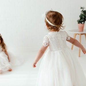 May include: Two young girls wearing white lace dresses and flower crowns. One girl is sitting on the floor while the other girl is standing and looking at her.