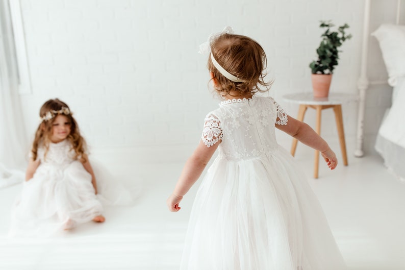 May include: Two young girls wearing white lace dresses and flower crowns. One girl is sitting on the floor while the other girl is standing and looking at her.