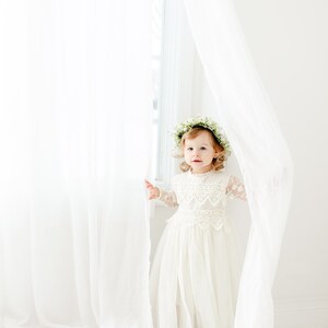 May include: A toddler wearing a cream-colored dress with long lace sleeves, a lace bodice overlay, and a full tulle skirt. The toddler is adorned with a delicate floral crown made of small white flowers and green leaves, standing between sheer white curtains.