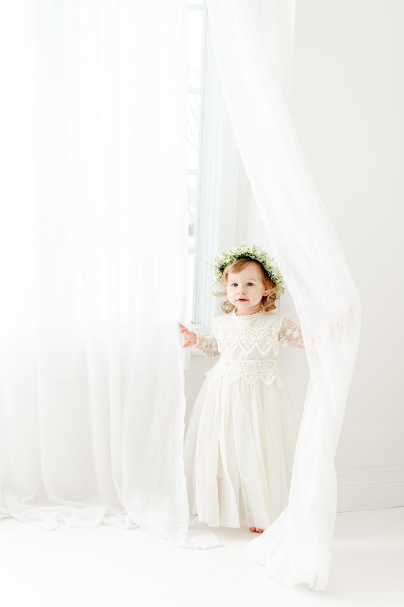 May include: A toddler wearing a cream-colored dress with long lace sleeves, a lace bodice overlay, and a full tulle skirt. The toddler is adorned with a delicate floral crown made of small white flowers and green leaves, standing between sheer white curtains.