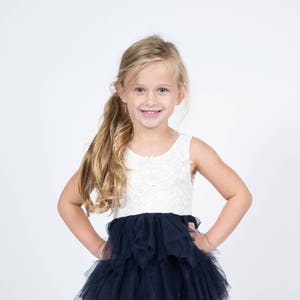 Puede incluir: Una ni&ntilde;a joven con el pelo largo y rubio sonr&iacute;e mientras lleva un vestido blanco sin mangas con una falda de tul negra.