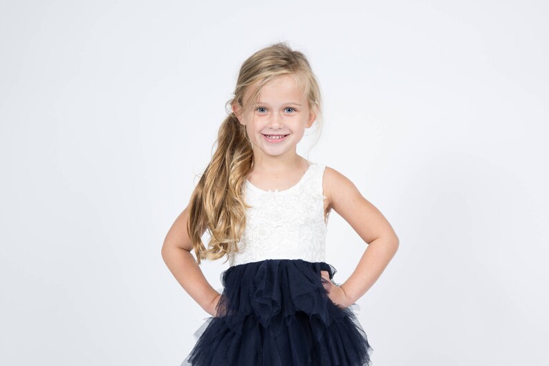 Puede incluir: Una ni&ntilde;a joven con el pelo largo y rubio sonr&iacute;e mientras lleva un vestido blanco sin mangas con una falda de tul negra.