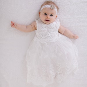 May include: A baby girl wearing a white lace dress and a white headband with a jeweled accent. She is lying on a white blanket and smiling.