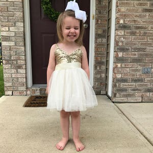 May include: A sleeveless dress featuring a gold sequined bodice with a V-neckline and a cream-colored tulle skirt. The dress includes a wide satin waistband and is paired with a large white hair bow accessory worn on the head.