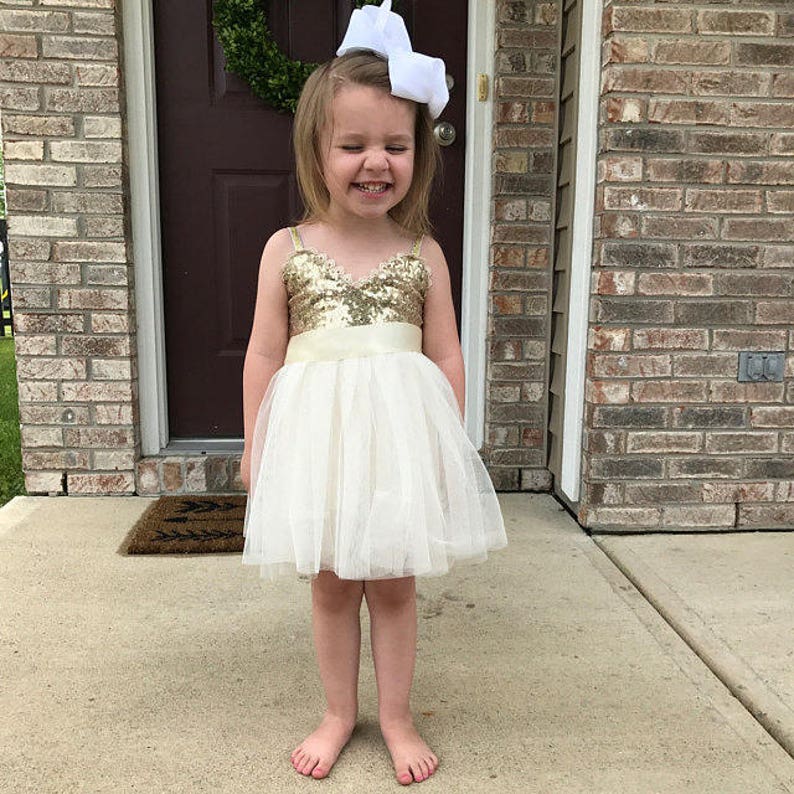 May include: A sleeveless dress featuring a gold sequined bodice with a V-neckline and a cream-colored tulle skirt. The dress includes a wide satin waistband and is paired with a large white hair bow accessory worn on the head.