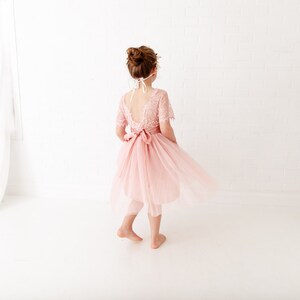 Pode incluir: Uma menina pequena usando um vestido de tule rosa claro com detalhes em renda e uma faixa de cetim. O vestido tem costas baixas e uma saia fluida.
