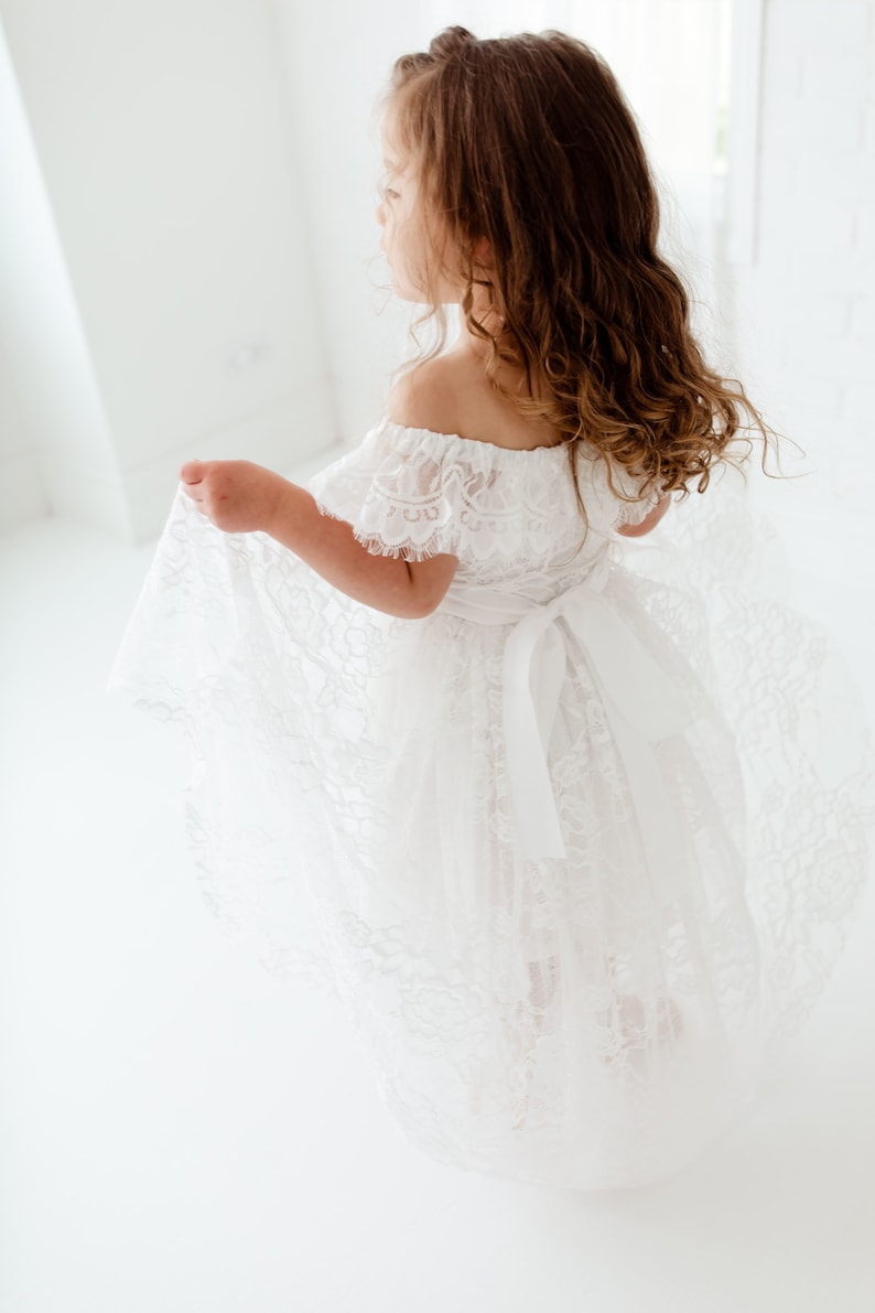 May include: A young girl wearing a white off-the-shoulder lace dress with a satin ribbon tied at the waist. The dress has a flowing skirt and is perfect for a special occasion.
