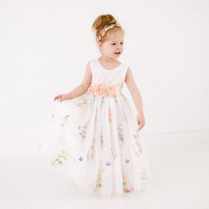 Puede incluir: Una ni&ntilde;a peque&ntilde;a con un vestido de tul blanco con estampado floral y una faja de flores rosa. Lleva una diadema dorada y tiene el pelo recogido en un mo&ntilde;o.