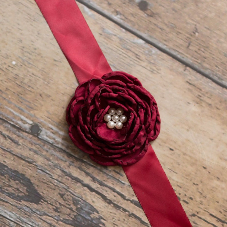 Peut inclure: Un ruban de satin bordeaux avec une grande fleur en tissu bordeaux orn&eacute;e de perles blanches.