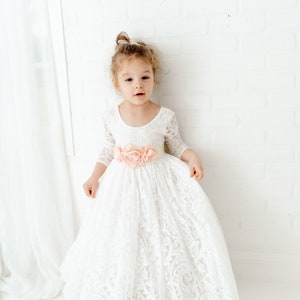 May include: A floor-length white lace dress with three-quarter length sleeves, a scoop neckline, and a wide waistband adorned with three large, soft peach-colored fabric flowers centered at the front of the garment's waistline.