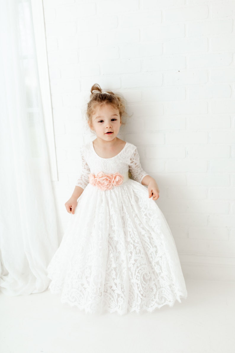 May include: A floor-length white lace dress with three-quarter length sleeves, a scoop neckline, and a wide waistband adorned with three large, soft peach-colored fabric flowers centered at the front of the garment's waistline.